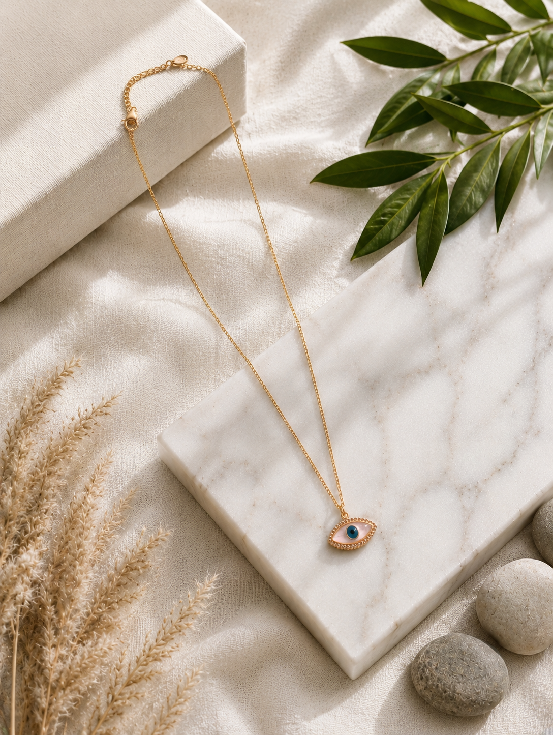 A gold chain necklace featuring an eye-shaped evil eye pendant with an iridescent pink mother-of-pearl inlay, a blue and black pupil, and a border of small sparkling crystals. The necklace in GN003_nazar.png is elegantly displayed on a square white marble block, surrounded by a textured linen cloth, dried pampas grass, smooth river stones, and a green leaf tip under warm, natural lighting.