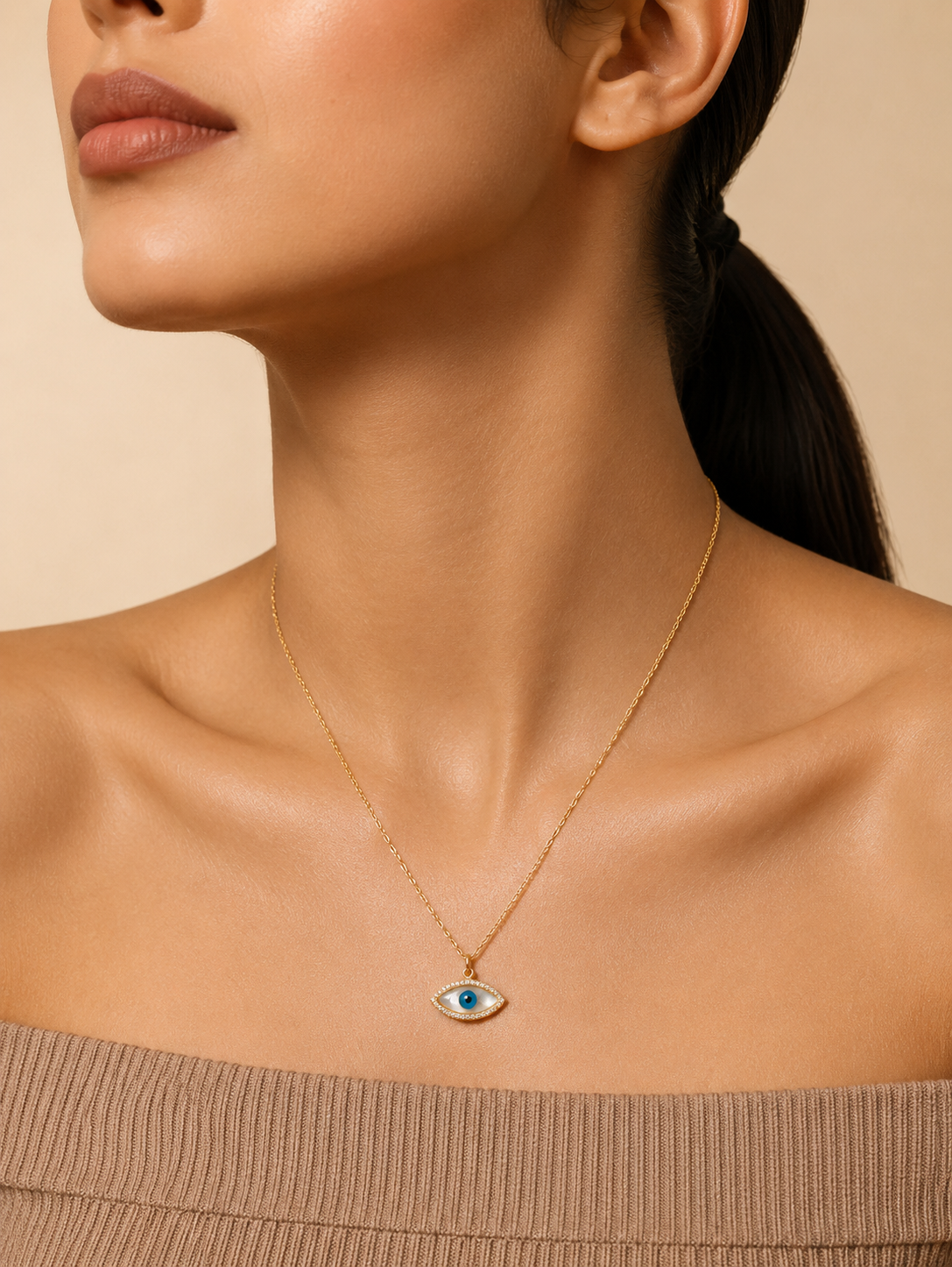 A gold chain necklace featuring an eye-shaped evil eye pendant with an iridescent pink mother-of-pearl inlay, a blue and black pupil, and a border of small sparkling crystals. The necklace in GN003_nazar.png is elegantly displayed on a square white marble block, surrounded by a textured linen cloth, dried pampas grass, smooth river stones, and a green leaf tip under warm, natural lighting.