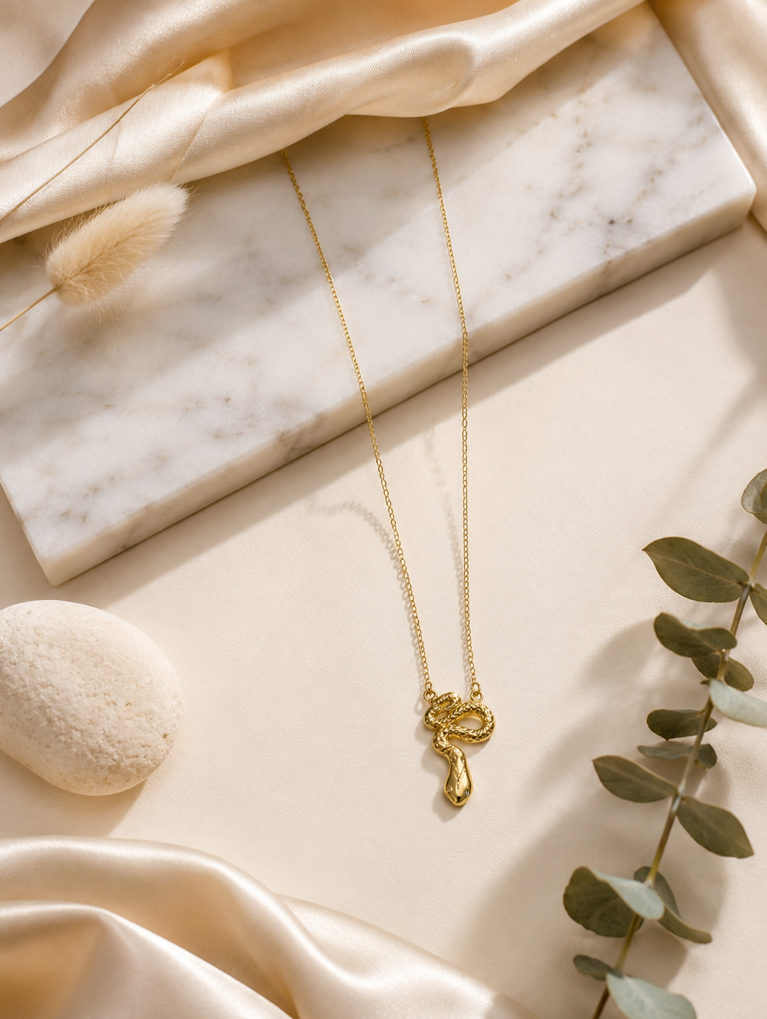 A fine gold chain necklace featuring a detailed, textured gold snake pendant shaped in a coiled slithering posture. In GN004_snake chain.png, the necklace hangs gracefully across a long white marble block, beautifully framed by shimmering champagne-colored silk fabric, a smooth cream-colored river stone, dried bunny tail grass, and a delicate sprig of eucalyptus under warm, naturally shadowed lighting.