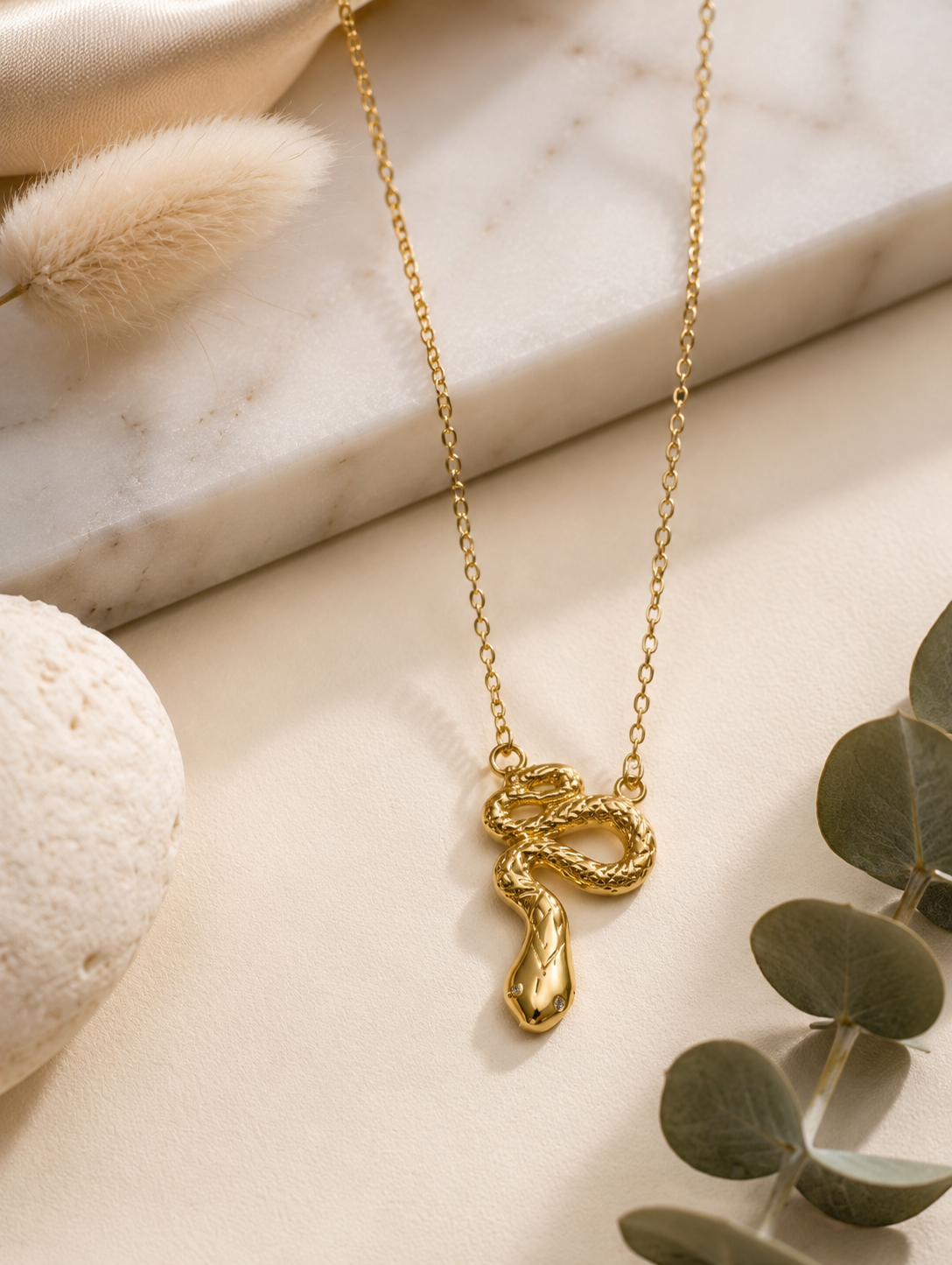 A fine gold chain necklace featuring a detailed, textured gold snake pendant shaped in a coiled slithering posture. In GN004_snake chain.png, the necklace hangs gracefully across a long white marble block, beautifully framed by shimmering champagne-colored silk fabric, a smooth cream-colored river stone, dried bunny tail grass, and a delicate sprig of eucalyptus under warm, naturally shadowed lighting.