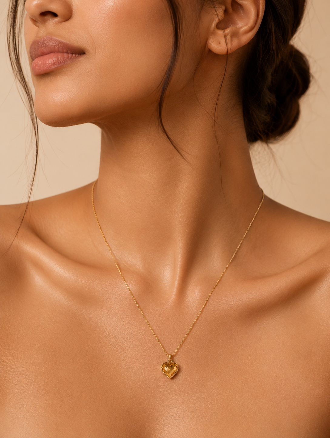 A close-up lifestyle shot in GN005_lilly chain_a.png showing a woman's collarbone and lower face as she wears a delicate gold necklace. The necklace features a small, faceted honey-amber crystal pendant shaped like a heart, enclosed in a textured gold border. The scene is illuminated by warm, soft lighting that highlights her skin and the glowing gemstone, with her hair styled up in a soft bun.