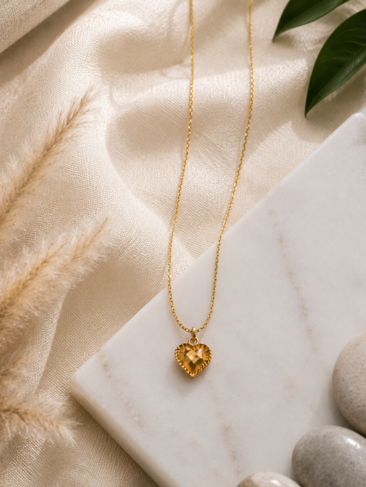 A close-up lifestyle shot in GN005_lilly chain_a.png showing a woman's collarbone and lower face as she wears a delicate gold necklace. The necklace features a small, faceted honey-amber crystal pendant shaped like a heart, enclosed in a textured gold border. The scene is illuminated by warm, soft lighting that highlights her skin and the glowing gemstone, with her hair styled up in a soft bun.