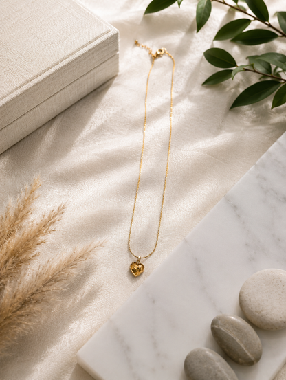 A close-up lifestyle shot in GN005_lilly chain_a.png showing a woman's collarbone and lower face as she wears a delicate gold necklace. The necklace features a small, faceted honey-amber crystal pendant shaped like a heart, enclosed in a textured gold border. The scene is illuminated by warm, soft lighting that highlights her skin and the glowing gemstone, with her hair styled up in a soft bun.