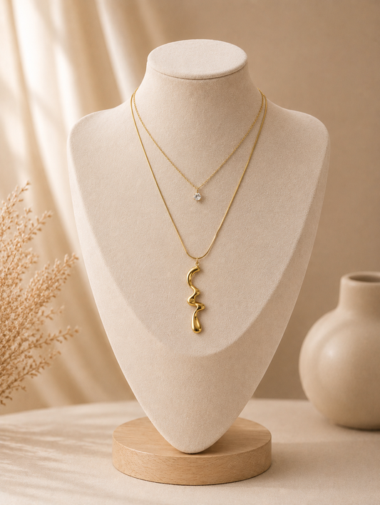 A multi-layered gold necklace displayed on a beige fabric mannequin bust. The necklace featured in GN006_double layered chain.png consists of two tiers: a shorter fine chain with a small round clear crystal solitaire pendant, and a longer chain with a wavy, organic abstract gold pendant. The studio background matches the warm, neutral color palette, featuring a ceramic vase, dried flora, and soft drapery under gentle light.