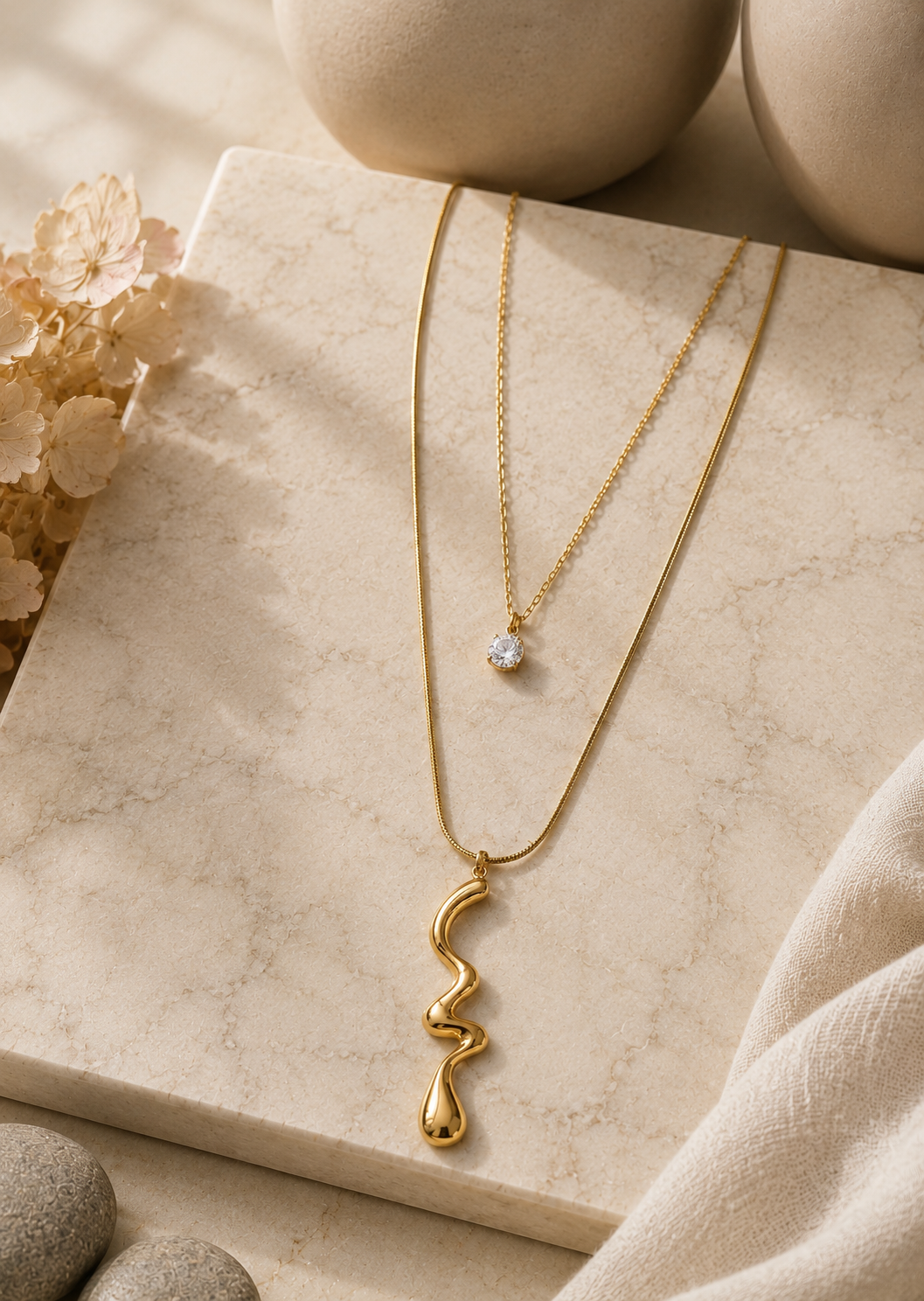 A multi-layered gold necklace displayed on a beige fabric mannequin bust. The necklace featured in GN006_double layered chain.png consists of two tiers: a shorter fine chain with a small round clear crystal solitaire pendant, and a longer chain with a wavy, organic abstract gold pendant. The studio background matches the warm, neutral color palette, featuring a ceramic vase, dried flora, and soft drapery under gentle light.