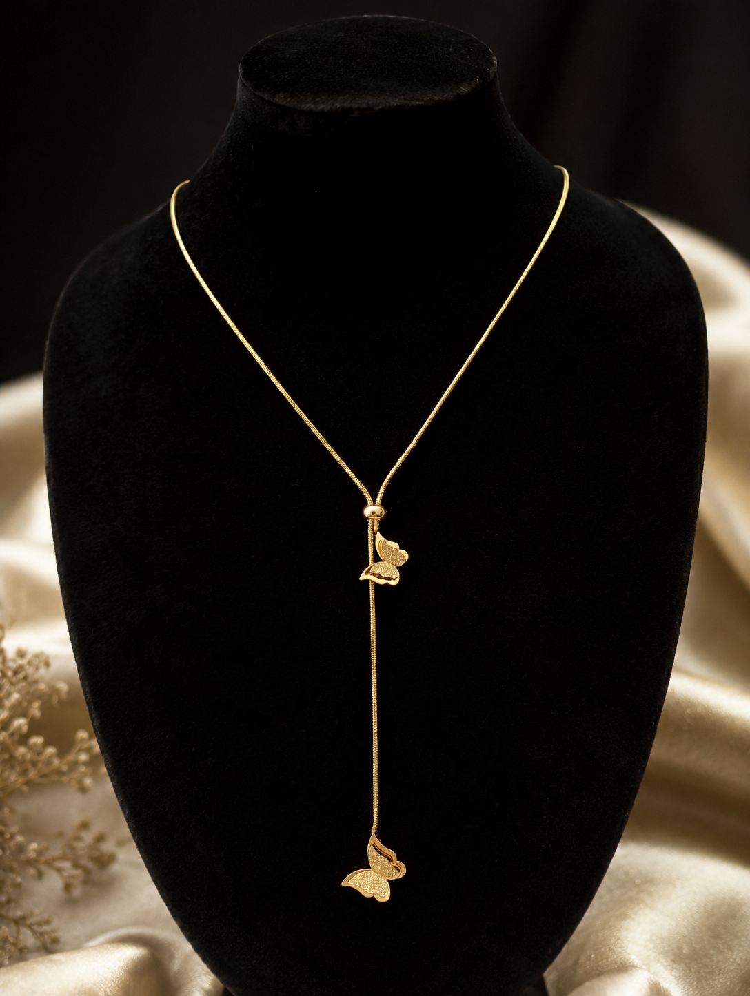A gold lariat-style necklace featuring two delicate butterfly charms attached to a drop chain with an adjustable sliding bead. In GN007_butterfly chain.png, the necklace is displayed on a long, veined white marble block. The luxury flat-lay arrangement is framed by shimmering, draped champagne silk fabric, a smooth cream river stone, fluffy dried bunny tail grass, and a sprig of green eucalyptus under warm, bright lighting.