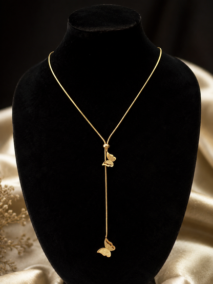 A gold lariat-style necklace featuring two delicate butterfly charms attached to a drop chain with an adjustable sliding bead. In GN007_butterfly chain.png, the necklace is displayed on a long, veined white marble block. The luxury flat-lay arrangement is framed by shimmering, draped champagne silk fabric, a smooth cream river stone, fluffy dried bunny tail grass, and a sprig of green eucalyptus under warm, bright lighting.