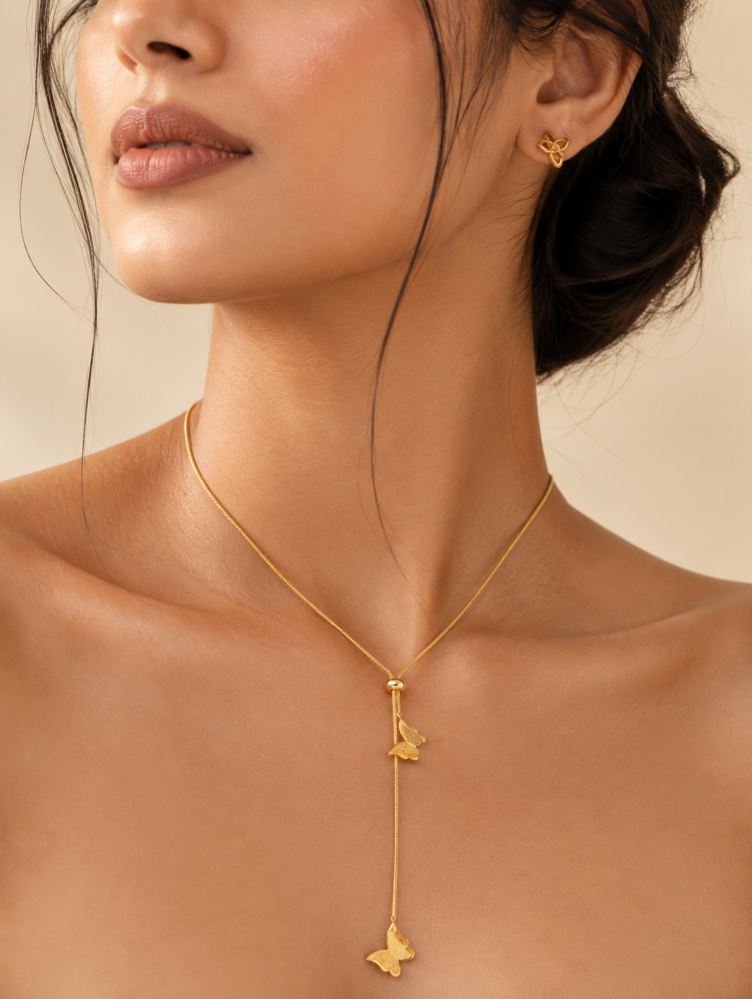 A gold lariat-style necklace featuring two delicate butterfly charms attached to a drop chain with an adjustable sliding bead. In GN007_butterfly chain.png, the necklace is displayed on a long, veined white marble block. The luxury flat-lay arrangement is framed by shimmering, draped champagne silk fabric, a smooth cream river stone, fluffy dried bunny tail grass, and a sprig of green eucalyptus under warm, bright lighting.