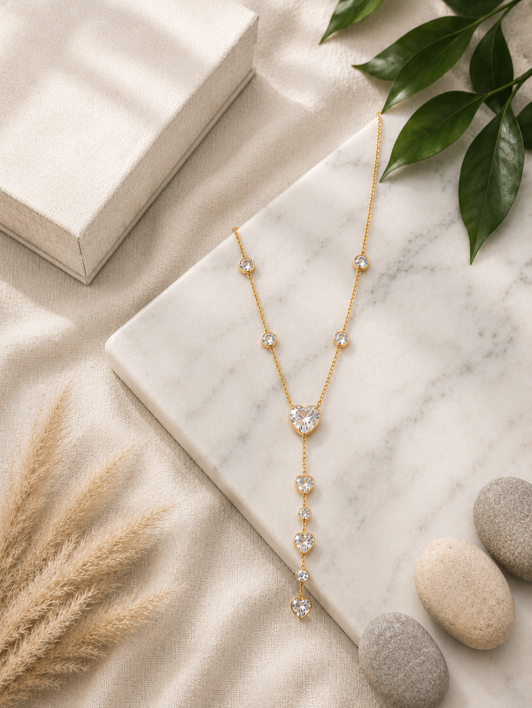 A fine gold lariat necklace featuring a cascading drop of faceted pink crystal hearts and small round bezel-set pink gems. In GN008_heart amora chain.png, the jewelry pieces drape beautifully from a rectangular white marble block across a beige tabletop. The luxury flat-lay setting is completed by a smooth cream river stone, fluffy dried bunny tail grass, draped champagne silk fabric, and a delicate branch of green eucalyptus under soft, natural lighting.