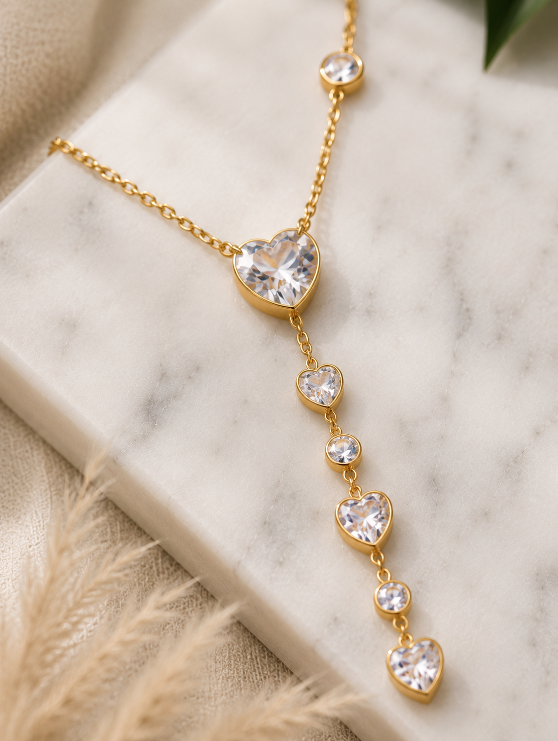 A fine gold lariat necklace featuring a cascading drop of faceted pink crystal hearts and small round bezel-set pink gems. In GN008_heart amora chain.png, the jewelry pieces drape beautifully from a rectangular white marble block across a beige tabletop. The luxury flat-lay setting is completed by a smooth cream river stone, fluffy dried bunny tail grass, draped champagne silk fabric, and a delicate branch of green eucalyptus under soft, natural lighting.