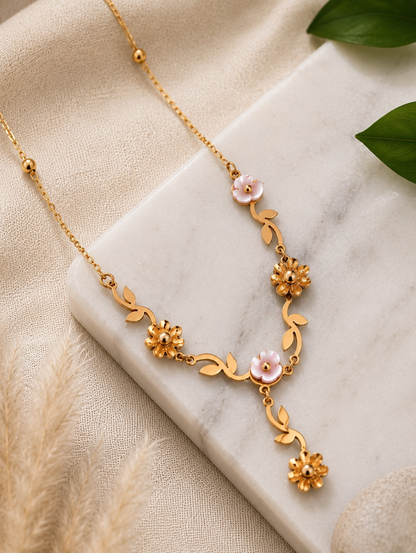 A close-up lifestyle shot in GN009_asymmetric floral chain_a.png showing a woman wearing a delicate, asymmetric gold necklace. The jewelry features an organic vine design decorated with small golden leaves, gold wildflowers, and white pearlescent blossoms, finished with a central dangling flower drop. The woman is wearing a ribbed, taupe off-the-shoulder top, and the scene is illuminated by soft, warm studio lighting that highlights her neckline and the necklace's intricate details.