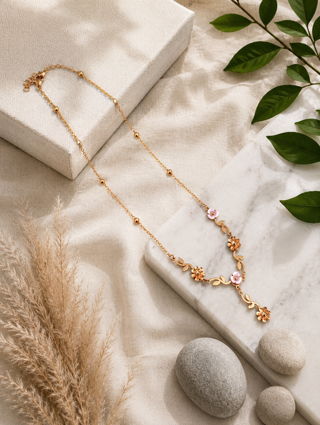 A close-up lifestyle shot in GN009_asymmetric floral chain_a.png showing a woman wearing a delicate, asymmetric gold necklace. The jewelry features an organic vine design decorated with small golden leaves, gold wildflowers, and white pearlescent blossoms, finished with a central dangling flower drop. The woman is wearing a ribbed, taupe off-the-shoulder top, and the scene is illuminated by soft, warm studio lighting that highlights her neckline and the necklace's intricate details.