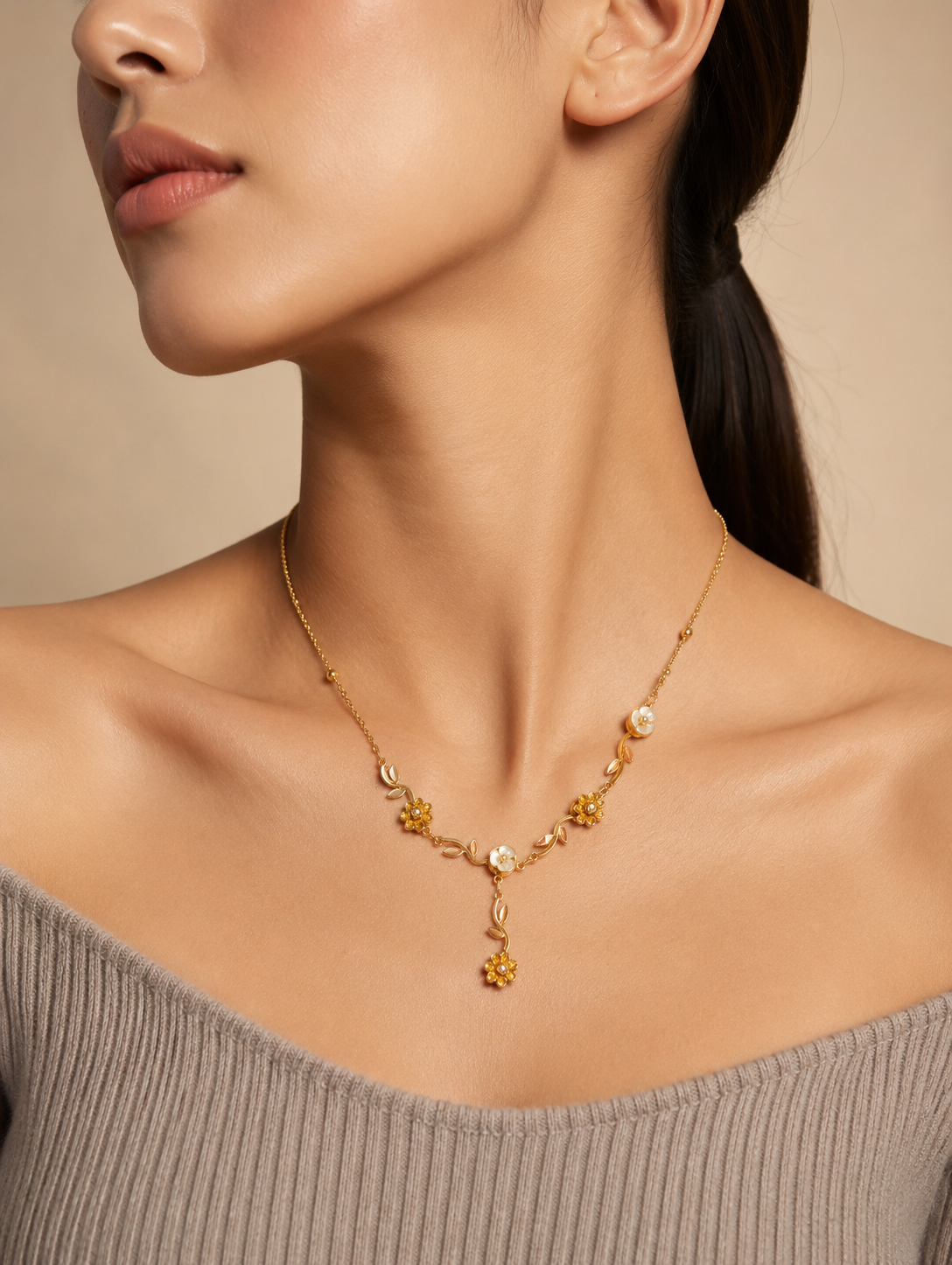 A close-up lifestyle shot in GN009_asymmetric floral chain_a.png showing a woman wearing a delicate, asymmetric gold necklace. The jewelry features an organic vine design decorated with small golden leaves, gold wildflowers, and white pearlescent blossoms, finished with a central dangling flower drop. The woman is wearing a ribbed, taupe off-the-shoulder top, and the scene is illuminated by soft, warm studio lighting that highlights her neckline and the necklace's intricate details.