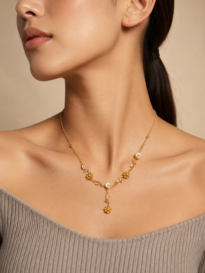 A close-up lifestyle shot in GN009_asymmetric floral chain_a.png showing a woman wearing a delicate, asymmetric gold necklace. The jewelry features an organic vine design decorated with small golden leaves, gold wildflowers, and white pearlescent blossoms, finished with a central dangling flower drop. The woman is wearing a ribbed, taupe off-the-shoulder top, and the scene is illuminated by soft, warm studio lighting that highlights her neckline and the necklace's intricate details.