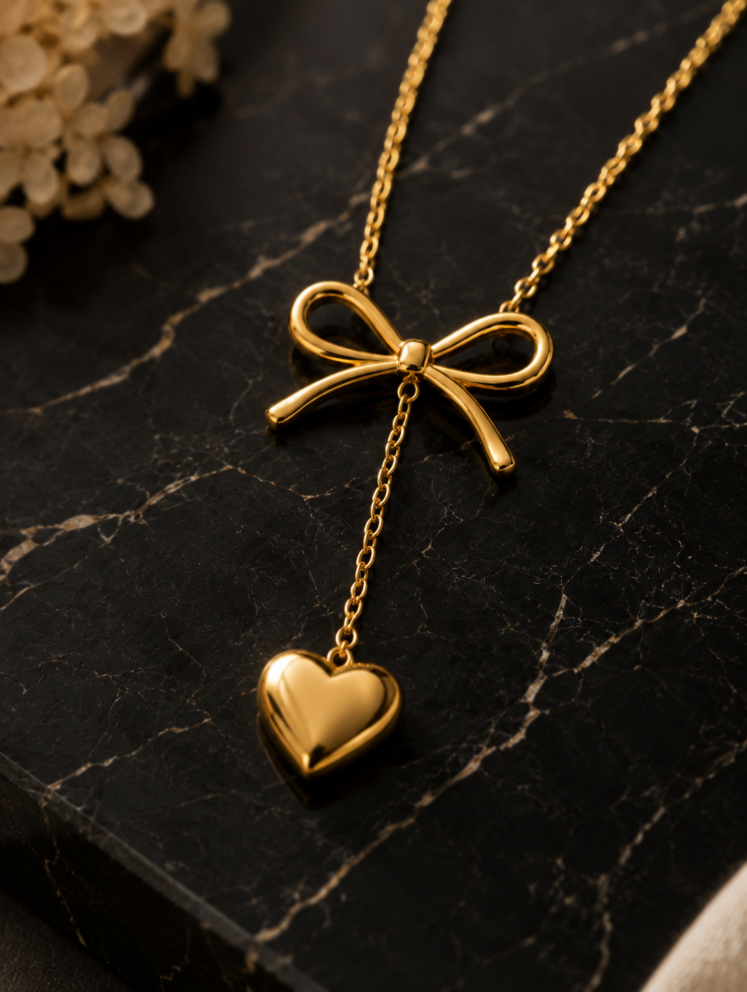 A detailed close-up shot of a high-polish gold necklace featured in GN010_asymmetric bow necklace_a.png. The necklace showcases a sleek, sculpted gold ribbon bow with a fine gold chain dropping from its center, ending in a smooth, puffed gold heart pendant. The jewelry is elegantly draped over a polished, dark black marble surface with striking white veins, illuminated by warm, atmospheric lighting with soft dried flowers blurred in the upper background.