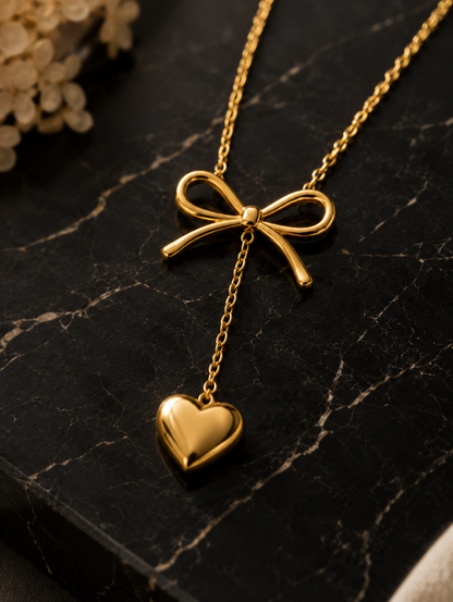 A detailed close-up shot of a high-polish gold necklace featured in GN010_asymmetric bow necklace_a.png. The necklace showcases a sleek, sculpted gold ribbon bow with a fine gold chain dropping from its center, ending in a smooth, puffed gold heart pendant. The jewelry is elegantly draped over a polished, dark black marble surface with striking white veins, illuminated by warm, atmospheric lighting with soft dried flowers blurred in the upper background.