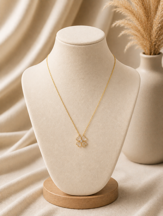 A delicate gold chain necklace with a four-leaf clover pendant displayed on a neutral beige fabric mannequin bust. The pendant in GN011_cute leaf chain.png features three heart-shaped leaves made of soft translucent stone, while the fourth leaf is filled with small sparkling crystals. The warm-toned, minimalist background includes draped soft fabric and a beige ceramic vase filled with dried pampas grass.