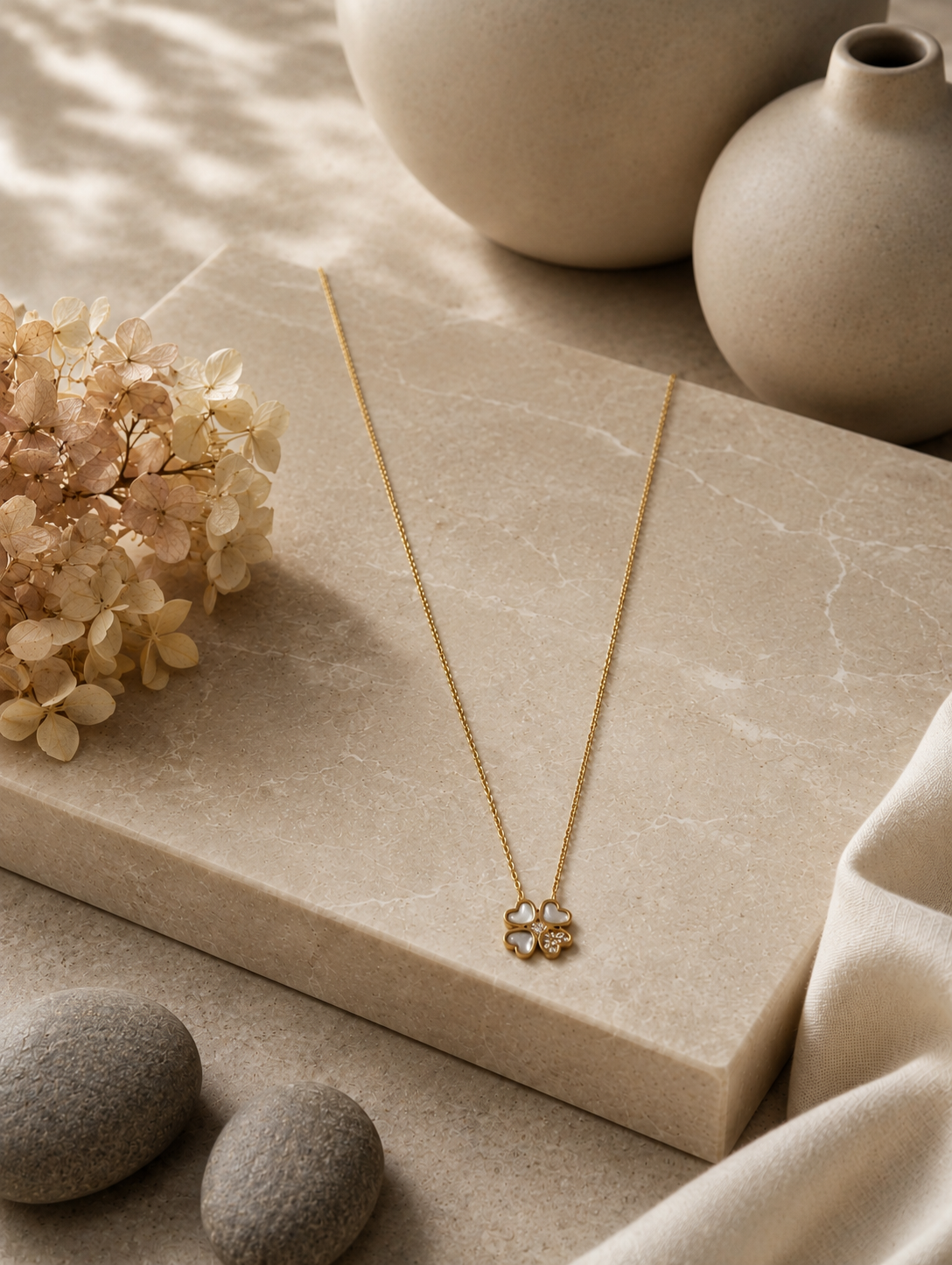 A delicate gold chain necklace with a four-leaf clover pendant displayed on a neutral beige fabric mannequin bust. The pendant in GN011_cute leaf chain.png features three heart-shaped leaves made of soft translucent stone, while the fourth leaf is filled with small sparkling crystals. The warm-toned, minimalist background includes draped soft fabric and a beige ceramic vase filled with dried pampas grass.