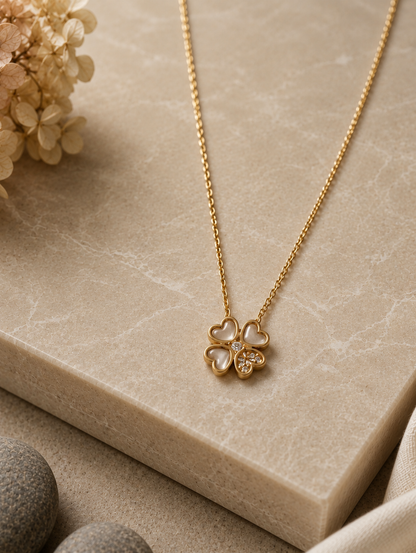 A delicate gold chain necklace with a four-leaf clover pendant displayed on a neutral beige fabric mannequin bust. The pendant in GN011_cute leaf chain.png features three heart-shaped leaves made of soft translucent stone, while the fourth leaf is filled with small sparkling crystals. The warm-toned, minimalist background includes draped soft fabric and a beige ceramic vase filled with dried pampas grass.