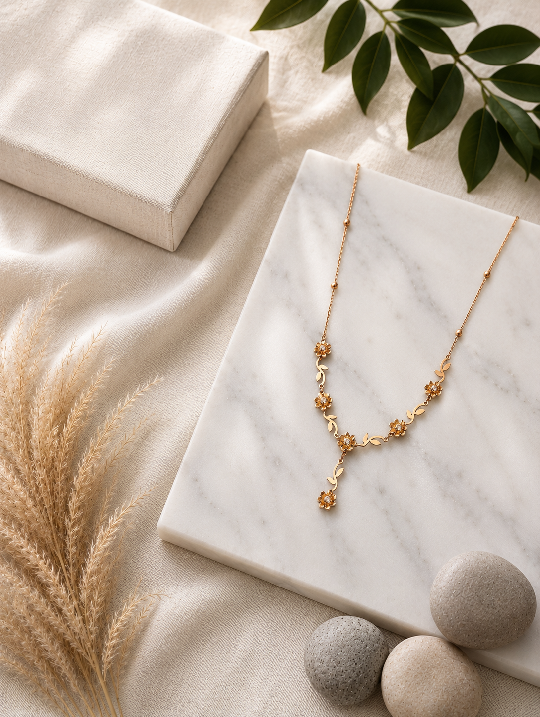A detailed flat-lay shot of an intricate gold necklace featuring a vine design with sculpted wildflowers, small golden leaves, and crystal-centered blossoms that culminate in a central dangling flower drop. In GN012_flower chain.png, the necklace is displayed across a white marble slab, set against a warm neutral background of wrinkled cream linen, a fabric-covered jewelry box, dried pampas grass, smooth textured river stones, and a sprig of fresh green leaves in the upper right corner.