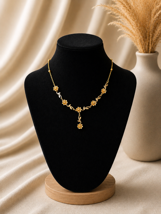 A detailed flat-lay shot of an intricate gold necklace featuring a vine design with sculpted wildflowers, small golden leaves, and crystal-centered blossoms that culminate in a central dangling flower drop. In GN012_flower chain.png, the necklace is displayed across a white marble slab, set against a warm neutral background of wrinkled cream linen, a fabric-covered jewelry box, dried pampas grass, smooth textured river stones, and a sprig of fresh green leaves in the upper right corner.