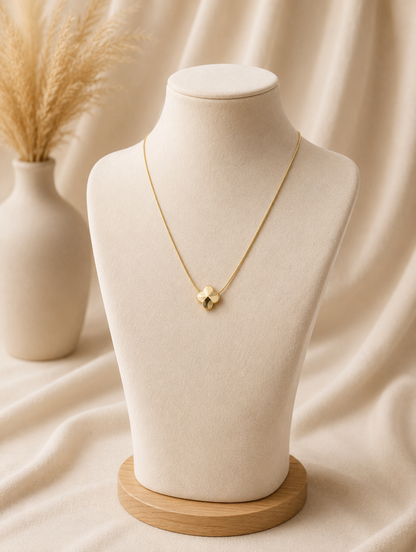 A minimalist gold pendant necklace featuring a polished four-leaf clover design, displayed on a beige fabric bust mannequin against a soft, neutral draped background with a vase of pampas grass. Stainless steel