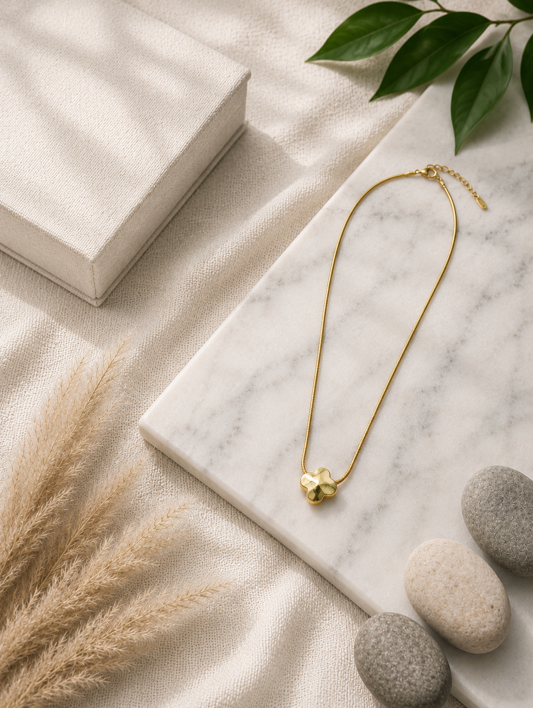 A minimalist gold pendant necklace featuring a polished four-leaf clover design, displayed on a beige fabric bust mannequin against a soft, neutral draped background with a vase of pampas grass. Stainless steel