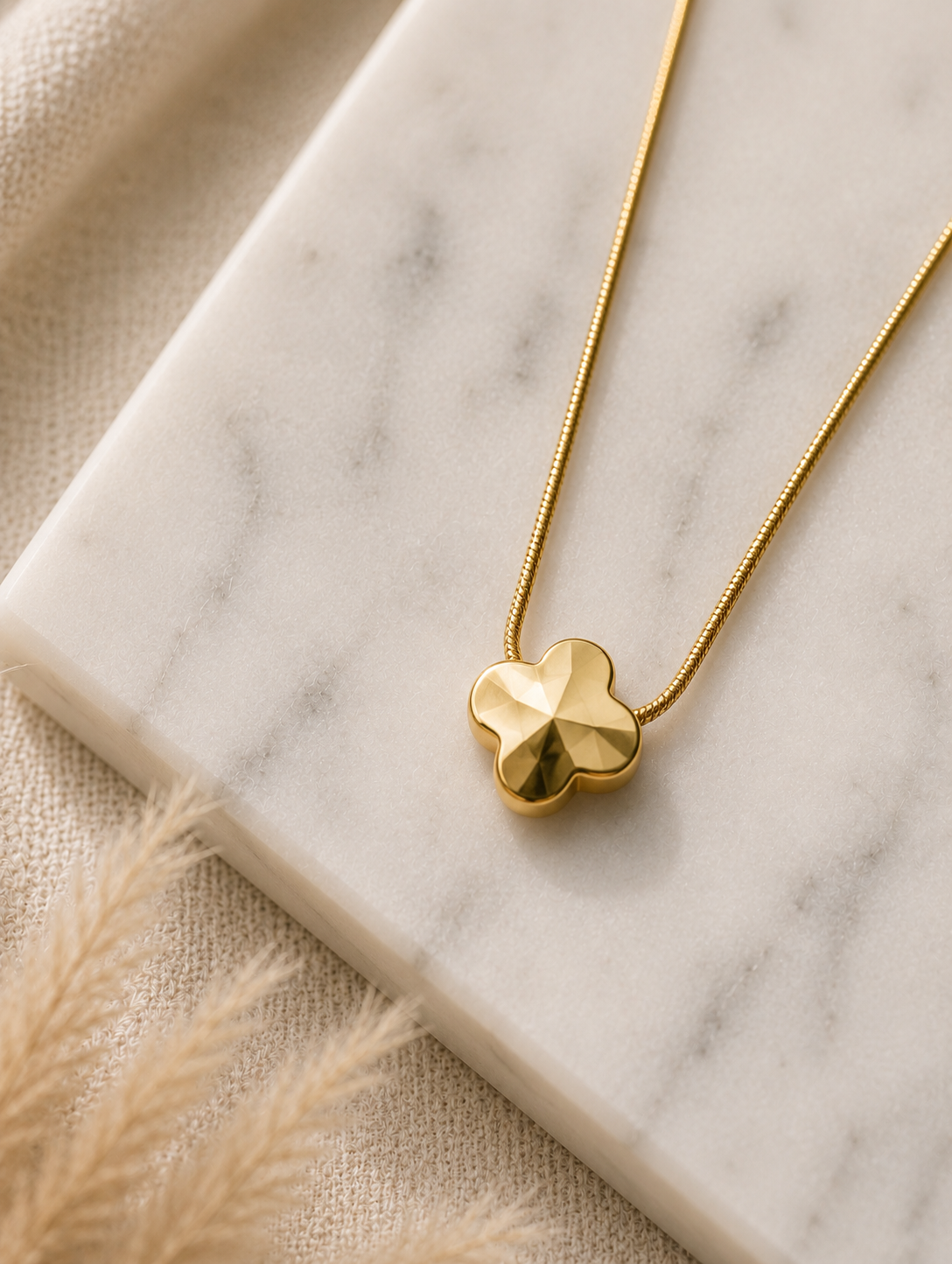 A minimalist gold pendant necklace featuring a polished four-leaf clover design, displayed on a beige fabric bust mannequin against a soft, neutral draped background with a vase of pampas grass. Stainless steel
