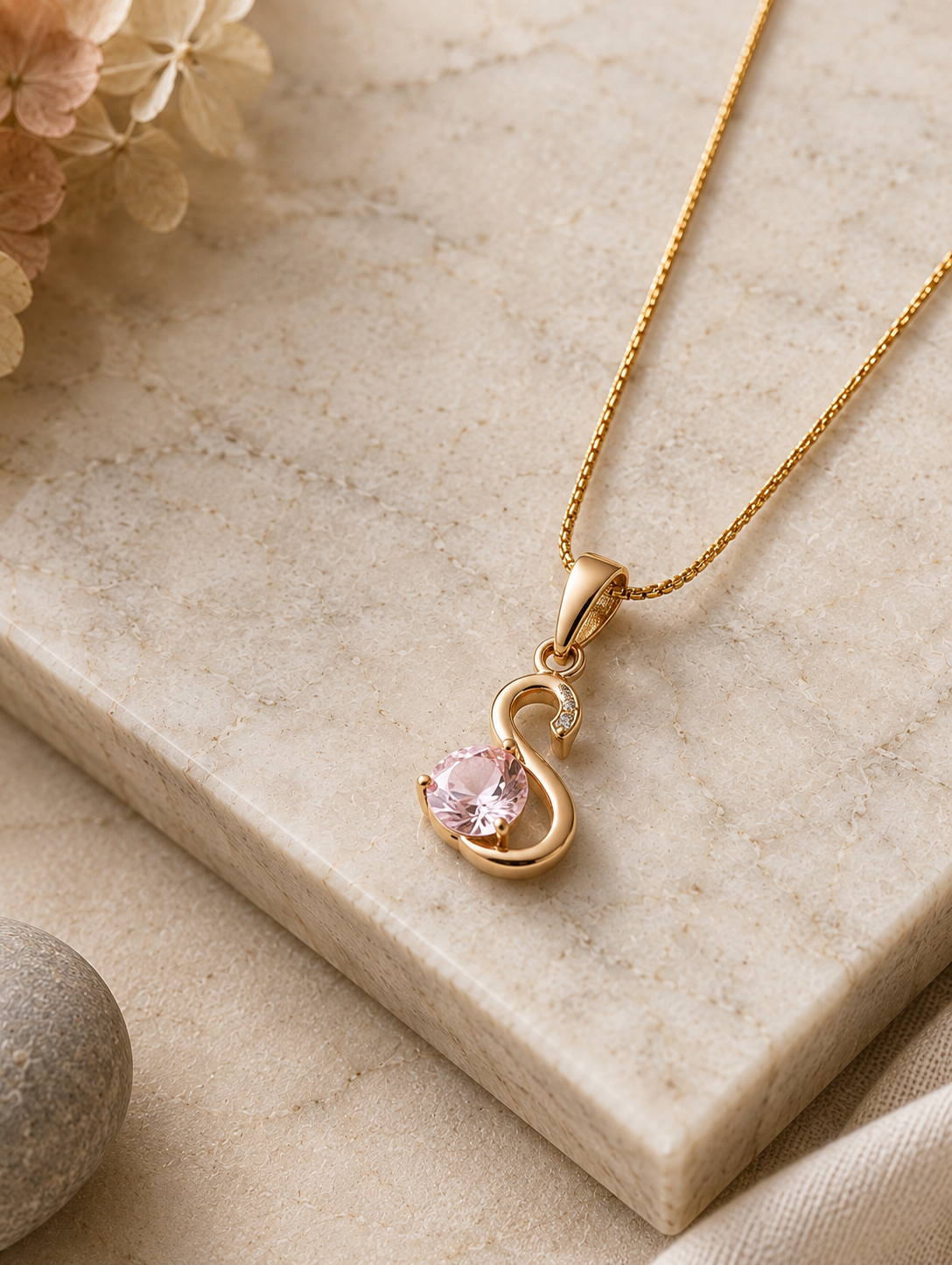 A  gold pendant necklace shaped like a stylized swan, featuring a faceted heart-shaped pink stone and a tiny clear crystal accent, displayed on a white marble slab with smooth stones and pampas grass in the background. Stainless Steel.
