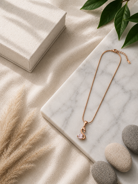 A  gold pendant necklace shaped like a stylized swan, featuring a faceted heart-shaped pink stone and a tiny clear crystal accent, displayed on a white marble slab with smooth stones and pampas grass in the background. Stainless Steel.