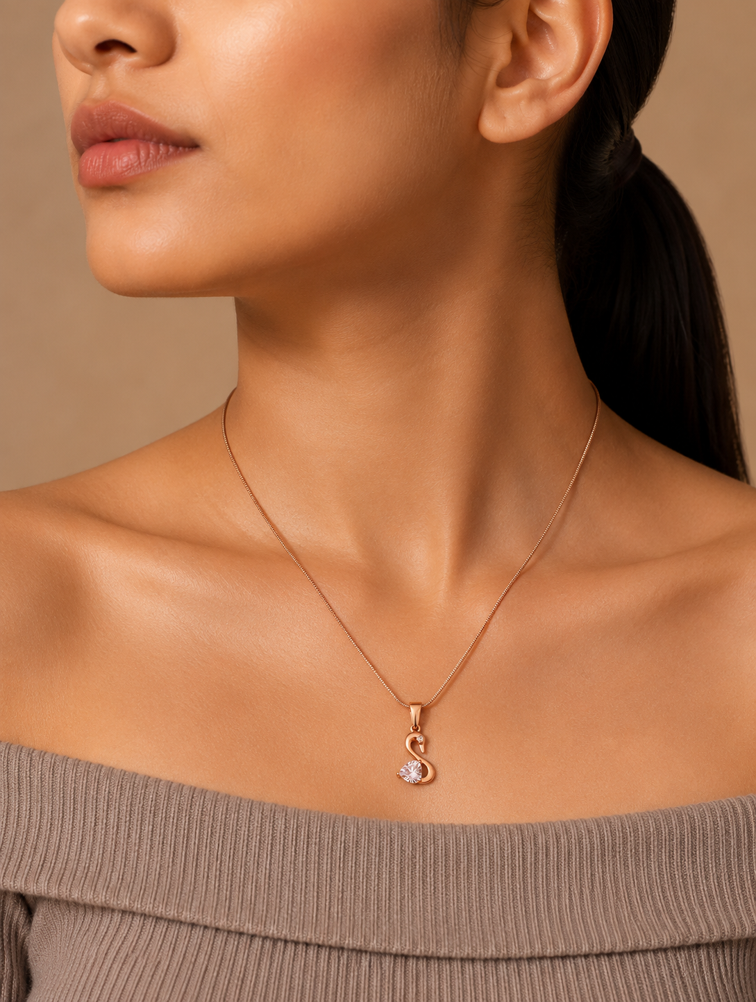 A  gold pendant necklace shaped like a stylized swan, featuring a faceted heart-shaped pink stone and a tiny clear crystal accent, displayed on a white marble slab with smooth stones and pampas grass in the background. Stainless Steel.