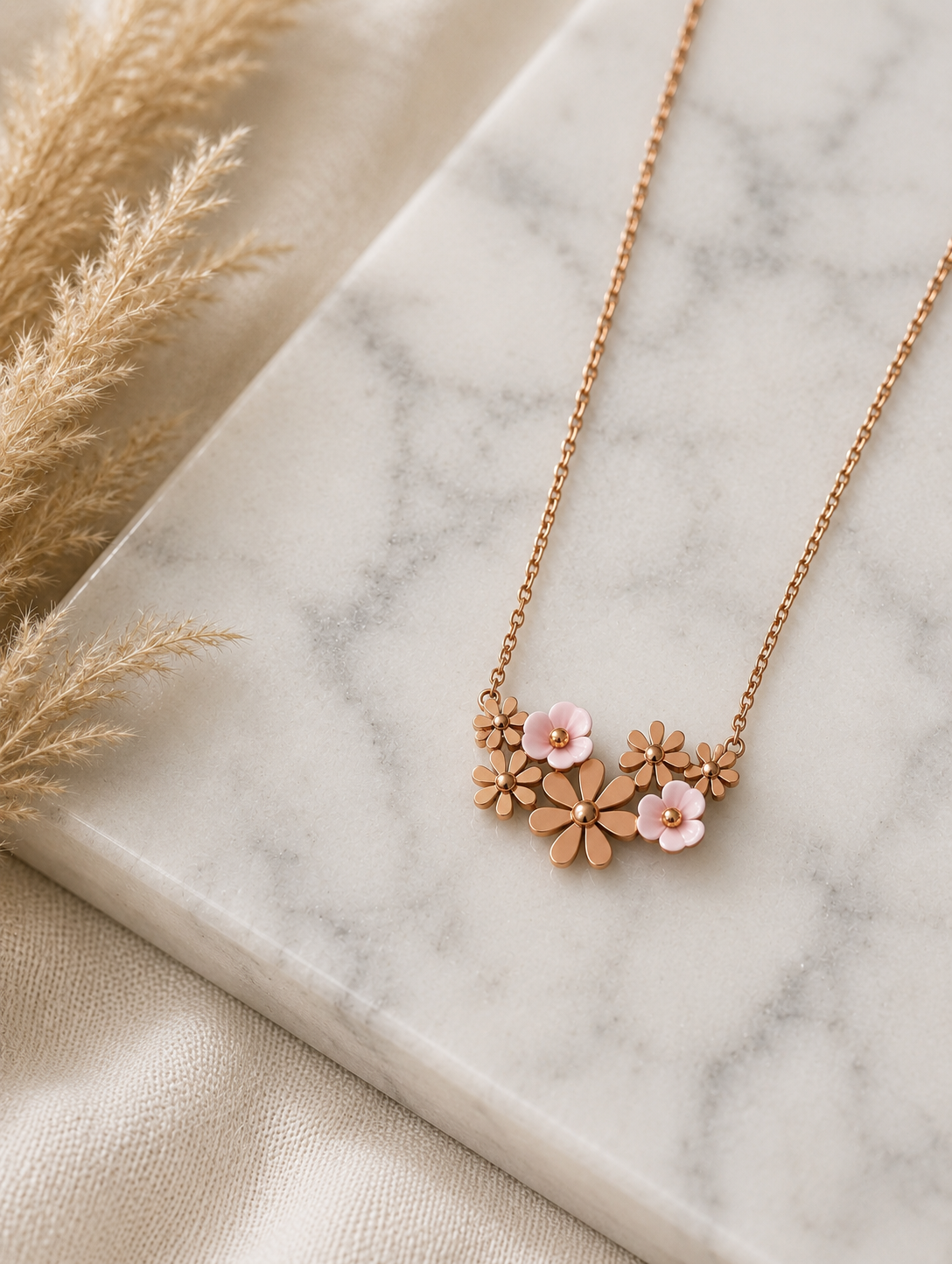 A gold necklace featuring an integrated cluster of small metallic flowers and soft pink enamel daisies, displayed diagonally across a white marble slab with dried pampas grass nearby. Stainless Steel.