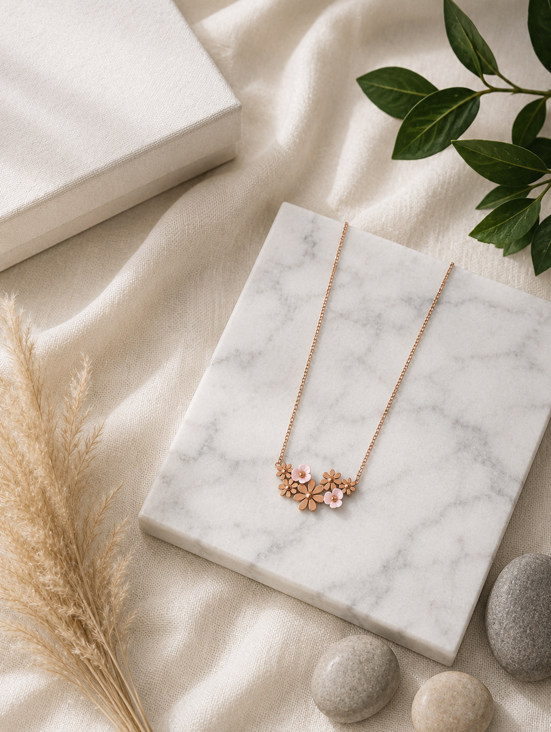 A gold necklace featuring an integrated cluster of small metallic flowers and soft pink enamel daisies, displayed diagonally across a white marble slab with dried pampas grass nearby. Stainless Steel.