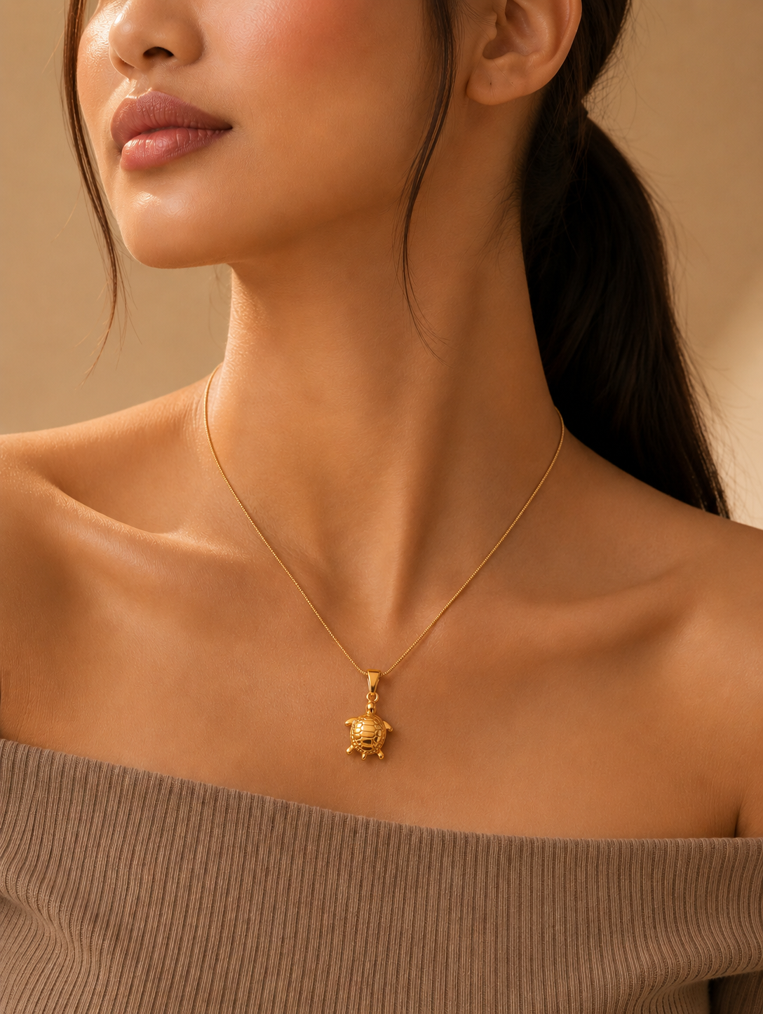 A close-up of a woman wearing a delicate gold necklace featuring a detailed turtle pendant with an engraved shell, draped elegantly against her collarbone while wearing a ribbed, off-the-shoulder brown top. Stainless Steel.