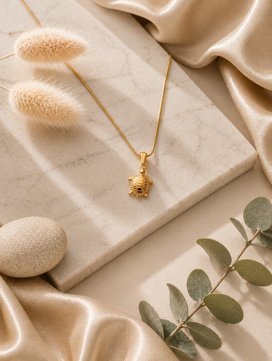 A close-up of a woman wearing a delicate gold necklace featuring a detailed turtle pendant with an engraved shell, draped elegantly against her collarbone while wearing a ribbed, off-the-shoulder brown top. Stainless Steel.