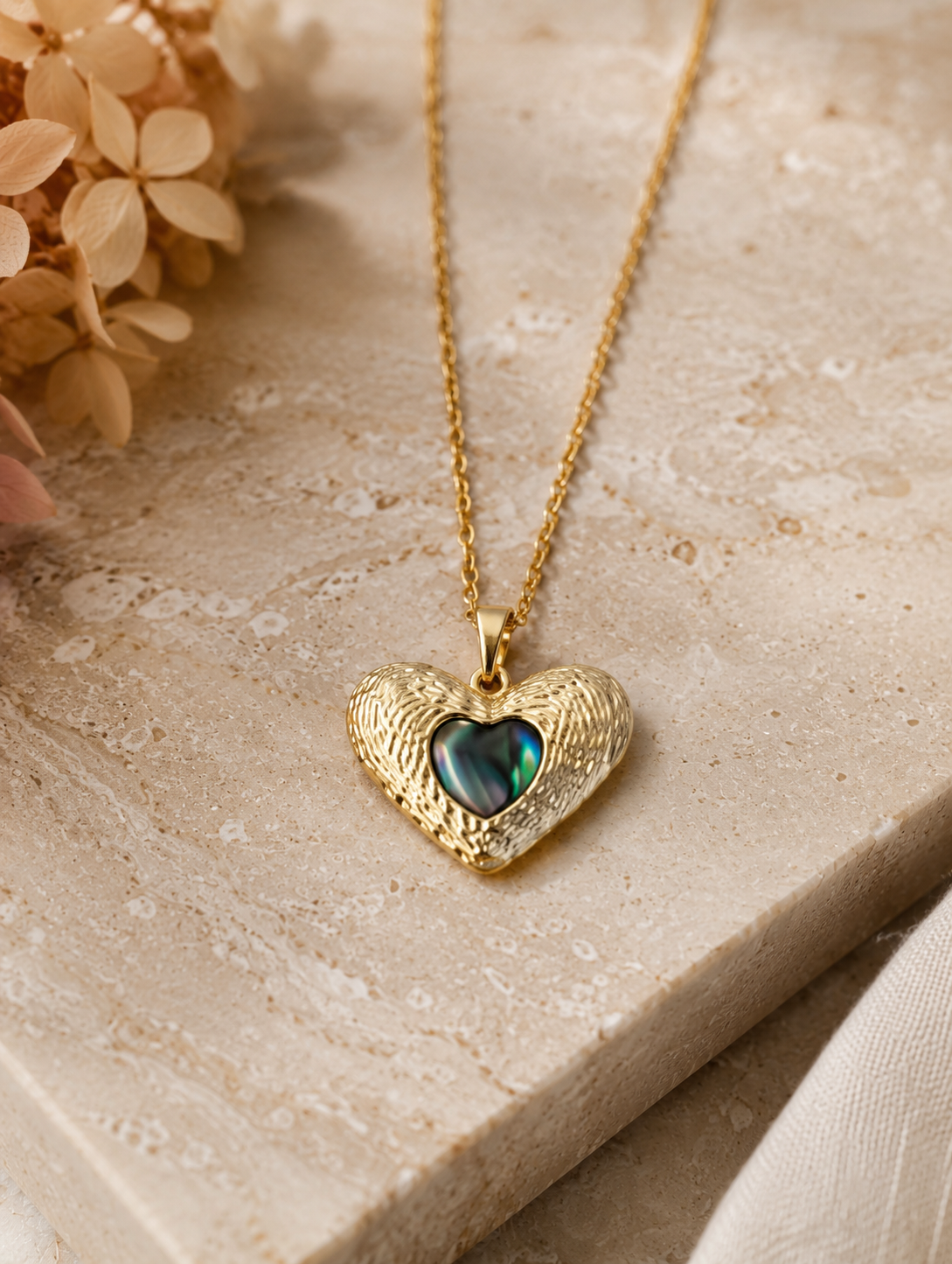 : A delicate gold-toned cable chain necklace featuring a contoured heart pendant with an intricate, ridged outer texture. The center of the pendant is inlaid with a smooth, glossy, iridescent heart that shifts between deep emerald green, teal, and black tones. The necklace is shown both as an elegant flat-lay display on a neutral marble stone block and worn by a model, showcasing its scale and placement on the neckline.