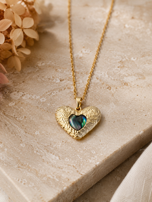 : A delicate gold-toned cable chain necklace featuring a contoured heart pendant with an intricate, ridged outer texture. The center of the pendant is inlaid with a smooth, glossy, iridescent heart that shifts between deep emerald green, teal, and black tones. The necklace is shown both as an elegant flat-lay display on a neutral marble stone block and worn by a model, showcasing its scale and placement on the neckline.
