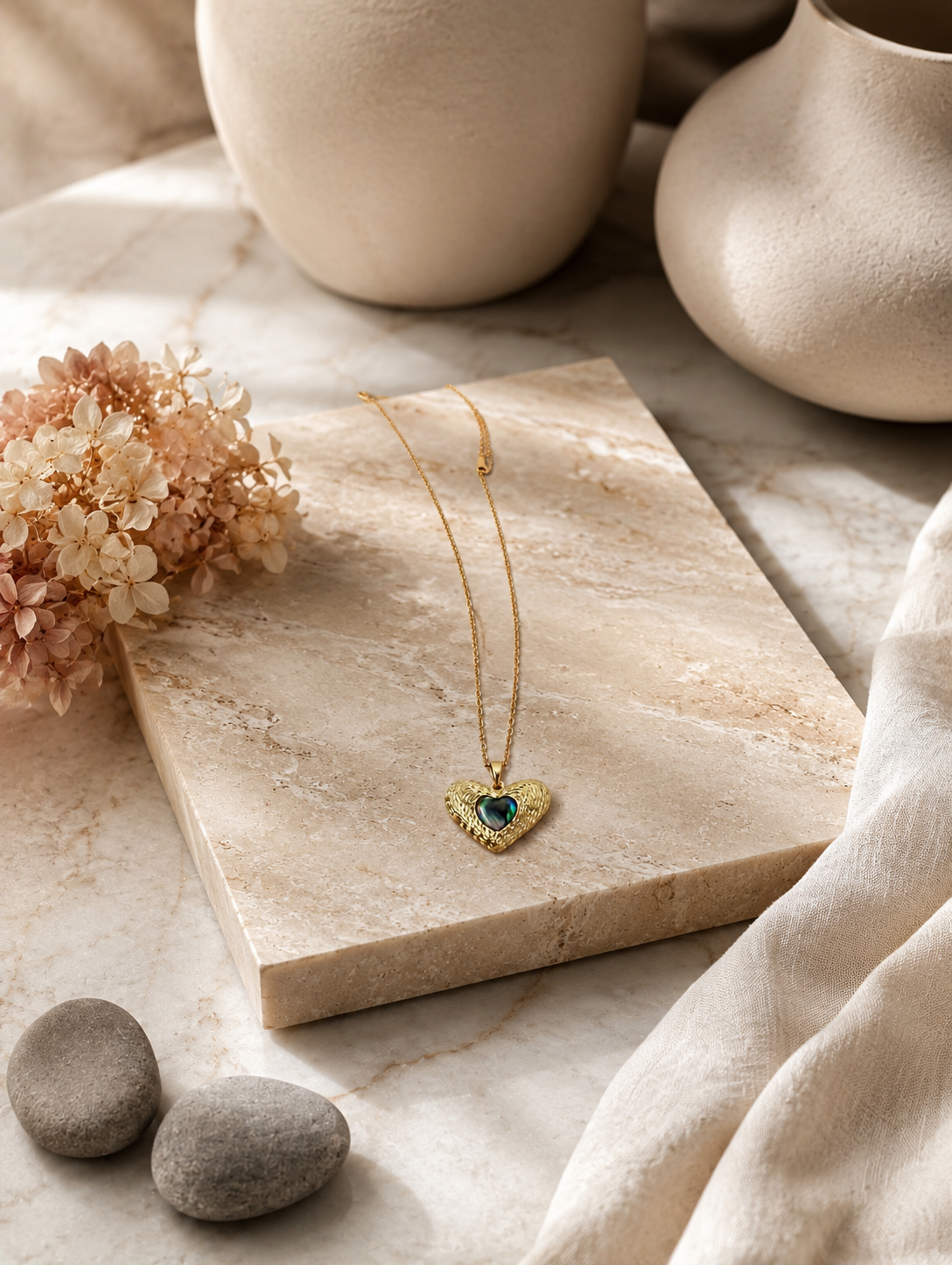 : A delicate gold-toned cable chain necklace featuring a contoured heart pendant with an intricate, ridged outer texture. The center of the pendant is inlaid with a smooth, glossy, iridescent heart that shifts between deep emerald green, teal, and black tones. The necklace is shown both as an elegant flat-lay display on a neutral marble stone block and worn by a model, showcasing its scale and placement on the neckline.