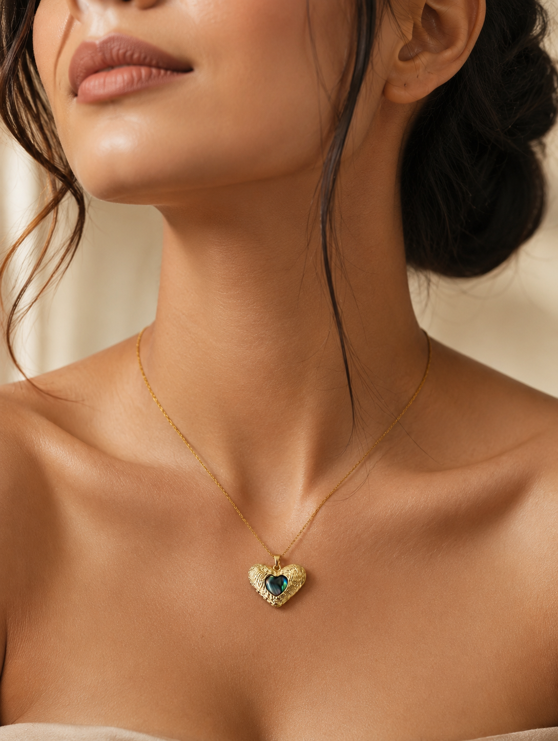 : A delicate gold-toned cable chain necklace featuring a contoured heart pendant with an intricate, ridged outer texture. The center of the pendant is inlaid with a smooth, glossy, iridescent heart that shifts between deep emerald green, teal, and black tones. The necklace is shown both as an elegant flat-lay display on a neutral marble stone block and worn by a model, showcasing its scale and placement on the neckline.