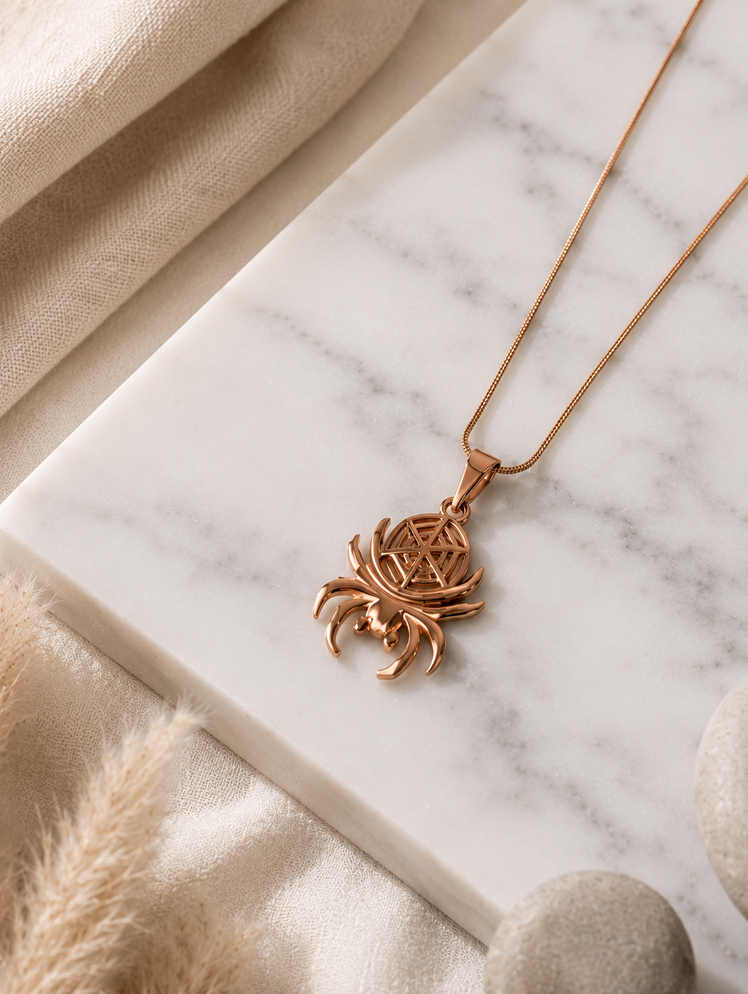 A polished rose gold-toned pendant necklace shaped like a spider, featuring curved legs and an intricate geometric web design cutout on the body. The pendant hangs from a thin, smooth snake chain and is displayed both as an aesthetic flat-lay on a white marble surface and draped over a neutral jewelry mannequin bust.