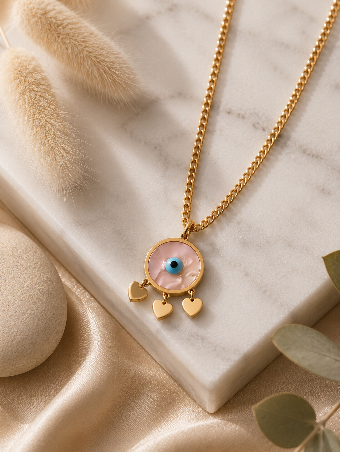 > **Alt Text:** A collection of product and lifestyle images featuring a gold-plated curb chain necklace with a circular evil eye pendant. The pendant showcases a pearlescent marbled inlay face, a vibrant cerulean blue and black eye at the center, and three dainty, polished gold heart charms dangling from its base. The necklace is displayed both as a flat-lay on a white marble slab with neutral aesthetic props—like a smooth river stone, dried grass, and draped champagne silk—and worn elegantly on a model's 