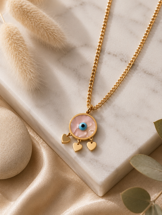 > **Alt Text:** A collection of product and lifestyle images featuring a gold-plated curb chain necklace with a circular evil eye pendant. The pendant showcases a pearlescent marbled inlay face, a vibrant cerulean blue and black eye at the center, and three dainty, polished gold heart charms dangling from its base. The necklace is displayed both as a flat-lay on a white marble slab with neutral aesthetic props—like a smooth river stone, dried grass, and draped champagne silk—and worn elegantly on a model's 