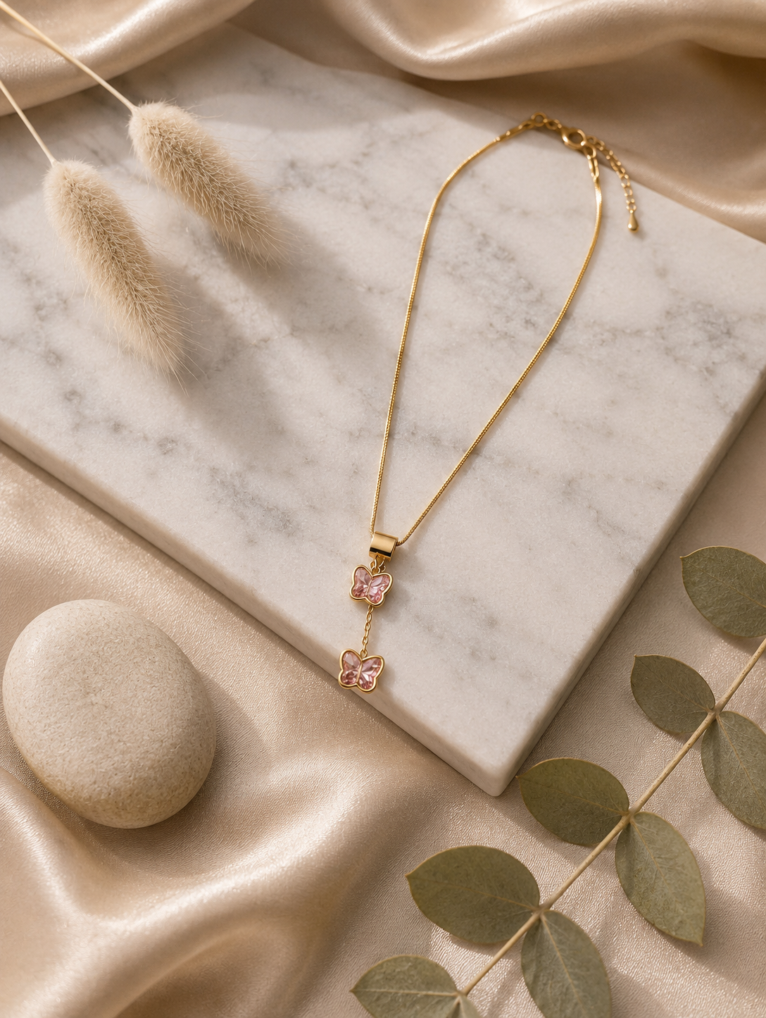 A collection of product and lifestyle images showcasing a gold-finished snake chain necklace with a unique tiered butterfly drop pendant. The pendant consists of two small butterflies stacked vertically, each crafted with polished gold borders and inlaid with faceted pink crystal wings. The top butterfly is attached to a wide, smooth gold bale, while the bottom butterfly hangs gently from a short drop chain. The necklace is pictured in elegant flat-lays on a white marble slab surrounded by neutral aesthetic