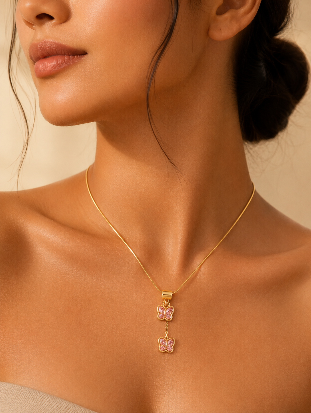 A collection of product and lifestyle images showcasing a gold-finished snake chain necklace with a unique tiered butterfly drop pendant. The pendant consists of two small butterflies stacked vertically, each crafted with polished gold borders and inlaid with faceted pink crystal wings. The top butterfly is attached to a wide, smooth gold bale, while the bottom butterfly hangs gently from a short drop chain. The necklace is pictured in elegant flat-lays on a white marble slab surrounded by neutral aesthetic