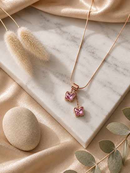 A collection of product and lifestyle images showcasing a gold-finished snake chain necklace with a unique tiered butterfly drop pendant. The pendant consists of two small butterflies stacked vertically, each crafted with polished gold borders and inlaid with faceted pink crystal wings. The top butterfly is attached to a wide, smooth gold bale, while the bottom butterfly hangs gently from a short drop chain. The necklace is pictured in elegant flat-lays on a white marble slab surrounded by neutral aesthetic