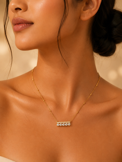 Alt Text: A close-up lifestyle photograph of a woman wearing a delicate gold-toned station chain necklace featuring a modern, horizontal bar pendant. The pendant consists of a thin metallic top bar encrusted with tiny sparkling crystals, directly underneath which is a row of five round-cut, brilliant clear crystals set side by side. The necklace rests elegantly on her collarbone against a softly lit, warm background.