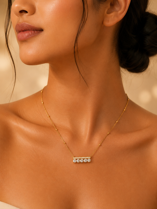 Alt Text: A close-up lifestyle photograph of a woman wearing a delicate gold-toned station chain necklace featuring a modern, horizontal bar pendant. The pendant consists of a thin metallic top bar encrusted with tiny sparkling crystals, directly underneath which is a row of five round-cut, brilliant clear crystals set side by side. The necklace rests elegantly on her collarbone against a softly lit, warm background.