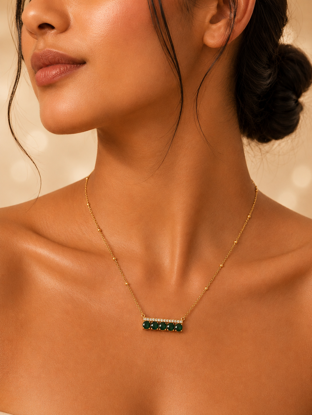 A close-up lifestyle photograph of a woman showcasing a gold-toned horizontal bar pendant necklace. The design features a thin top bar lined with a row of tiny, shimmering pavé-set crystals, with five round-cut, rich emerald-green stones arranged side by side below it. The pendant is suspended from a delicate chain accented with tiny gold beads, resting against the model's collarbone.