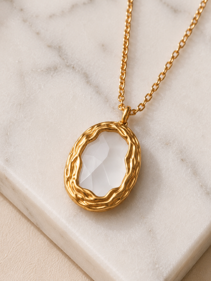 A close-up photograph from GN028_white milky pendent_399.png showing a woman wearing a delicate gold chain necklace with a vintage-inspired oval pendant, which features a smooth, milky-white stone framed by a textured, braided gold-tone border.