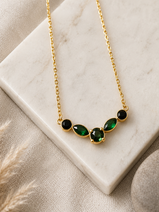 A product photography collage featuring a delicate gold-toned necklace with a curved station pendant of five faceted gemstones in rich emerald green and deep black. The first view shows a close-up of the necklace resting on a white marble slab with smooth stones and textured linen. The second shows the entire length of the gold link chain in a styled flat-lay next to a gift box and tropical leaves. The third shows the necklace being worn by a model, resting elegantly on her collarbone with a taupe off-the-s